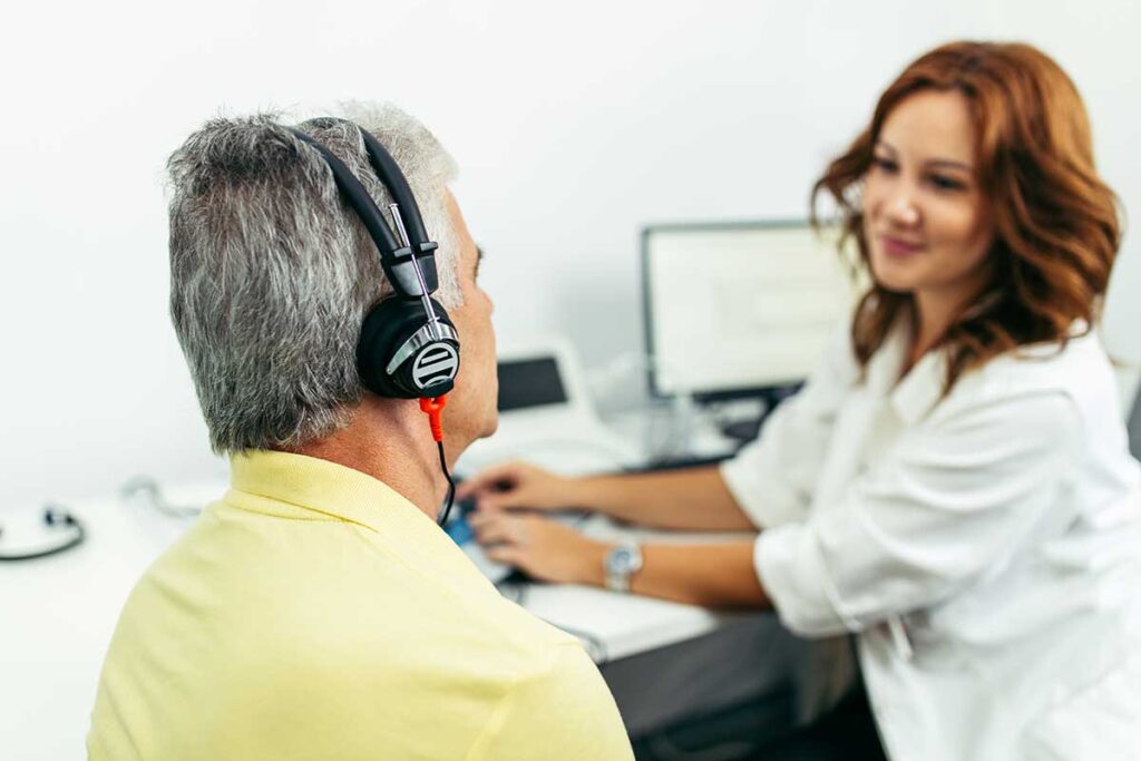 An audiologist demonstrates a hearing test with a local resident wearing headphones in a quiet exam room.
