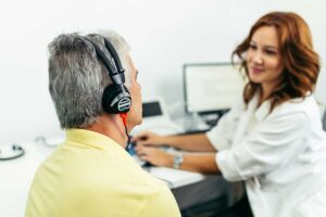 An audiologist demonstrates a hearing test with a local resident wearing headphones in a quiet exam room.